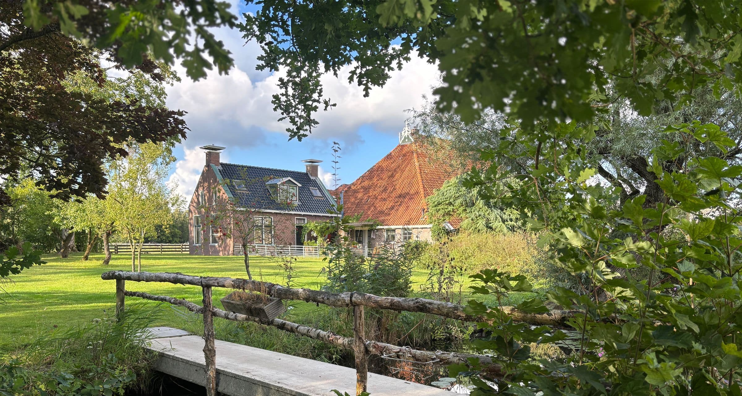 De boerderij van Hiddema State, gezien vanaf een houten bruggetje tussen de bomen.