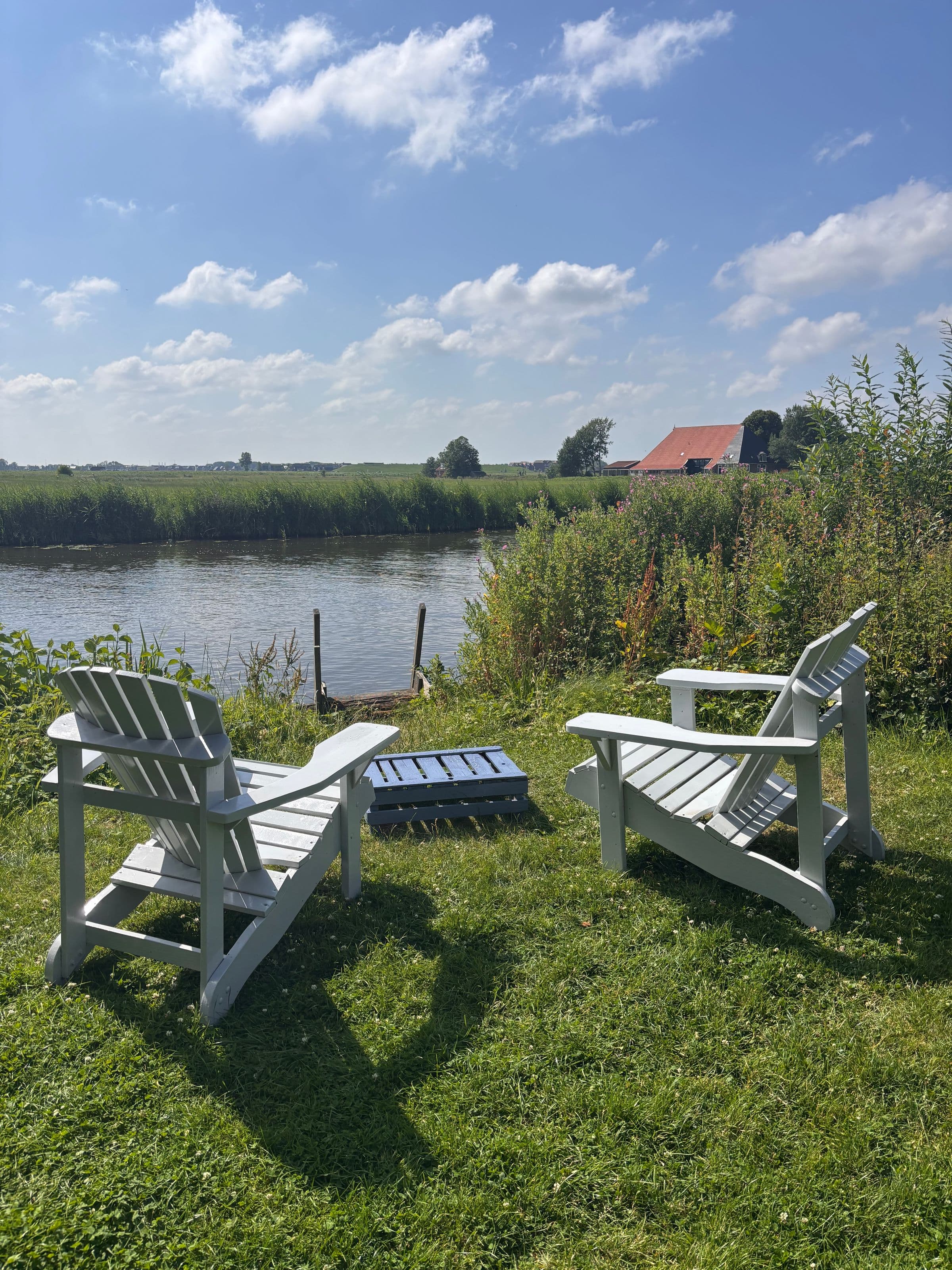 Twee witte tuinstoelen in het gras aan de vaart, met daarachter het open land en een boerderij.